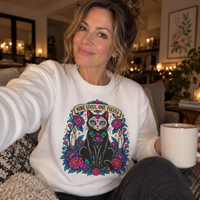 Load image into Gallery viewer, Woman in a white Nine Lives, One Fiesta Day of the Dead cat sweatshirt holding a mug in a cozy living room
