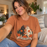Load image into Gallery viewer, Woman wearing the Love Has Nine Lives Day of the Dead black cat t-shirt in Yam, Comfort Colors, at home
